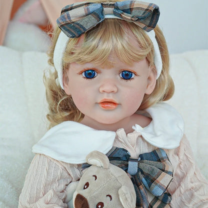  Close-up of reborn toddler doll with a plaid bow headband, dressed in a plaid dress with a white collar, holding a teddy bear.