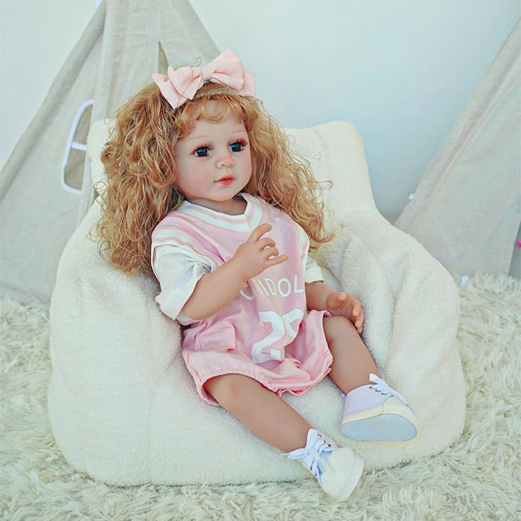 Reborn toddler doll sitting comfortably with a pink dress and bow, her legs extended on a soft surface.