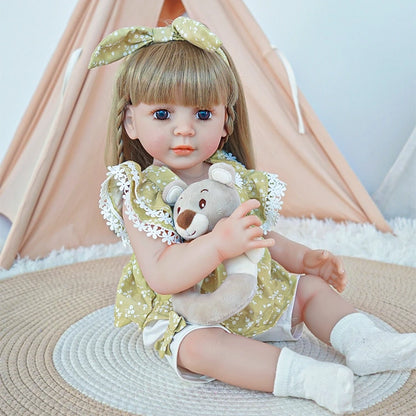 Reborn toddler doll sitting on a soft surface, wearing a green floral dress with lace accents, holding a teddy bear, looking gently to the side.