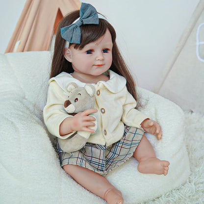 Reborn toddler doll sitting on a soft surface, wearing a cream-colored top with a plaid skirt, holding a teddy bear.