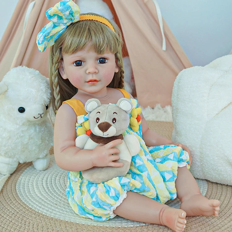 Reborn toddler doll sitting on a rug, wearing a yellow and blue outfit with a matching headband, holding a plush bear.