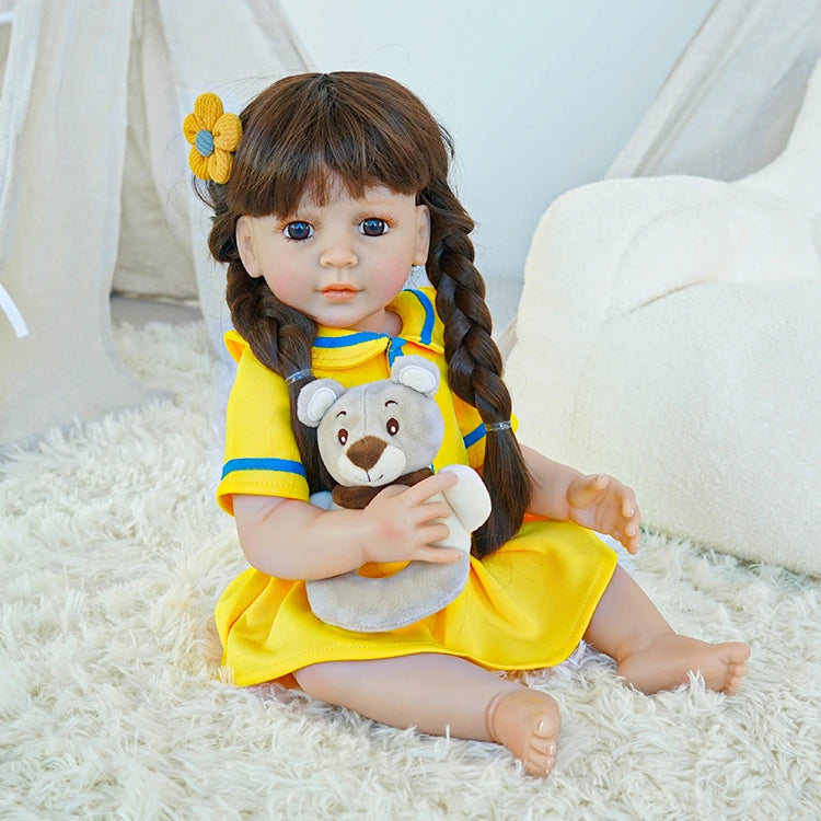 Reborn toddler doll sitting on a soft surface, wearing a yellow dress with blue accents and holding a teddy bear, looking gently to the side.