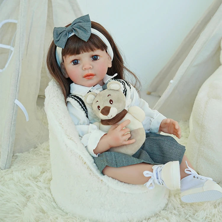 Reborn toddler doll sitting on a soft rug, wearing a gray and white school-themed outfit with a matching bow, holding a plush bear.