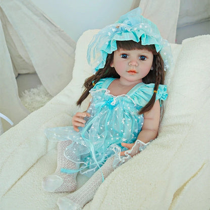 Reborn toddler doll sitting in a plush chair, wearing a blue dress and bonnet, with her hands resting on her lap.