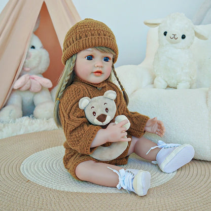 Reborn toddler doll sitting on a soft surface, dressed in a brown knit sweater and hat, holding a plush bear.