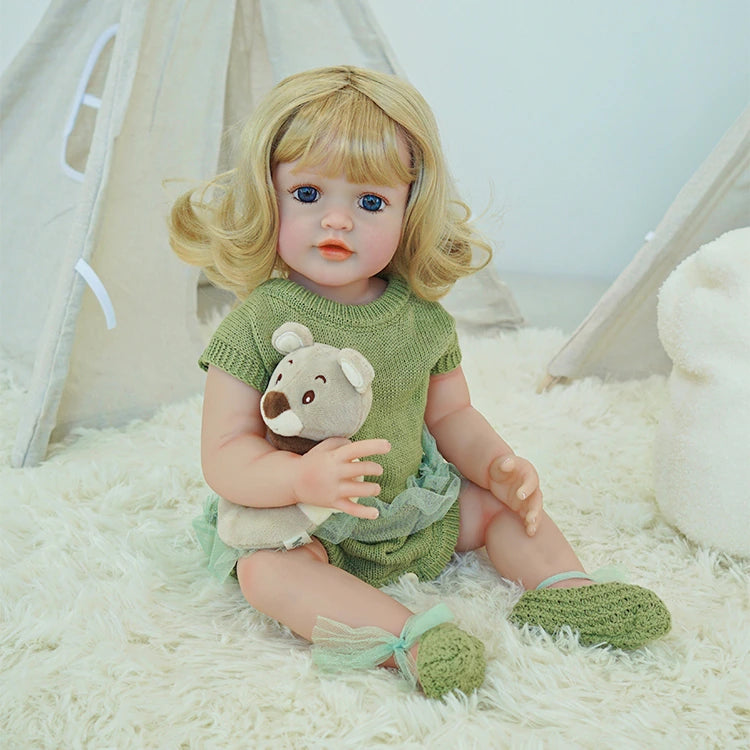  Reborn toddler doll sitting up with a teddy bear in her lap, dressed in a green outfit and matching shoes.