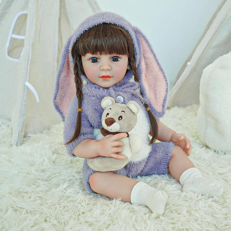 Reborn toddler doll in a purple bunny outfit, sitting on a plush rug and holding a teddy bear.