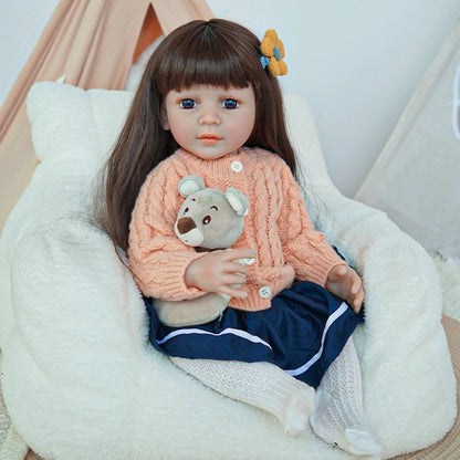 Reborn toddler doll sitting on a soft surface, wearing a peach sweater and navy skirt, holding a small teddy bear.