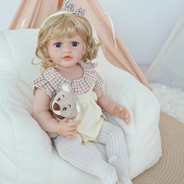 Reborn toddler doll sitting on a soft surface, wearing a pink sweater and white overalls, holding a teddy bear with a bunny face on the outfit.