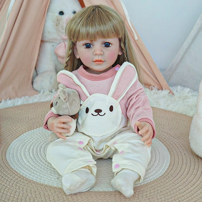 Reborn toddler doll sitting on a soft surface, wearing a pink sweater and white overalls, holding a teddy bear with a bunny face on the outfit.