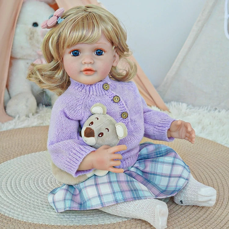 Reborn toddler doll sitting on a soft surface, wearing a purple sweater and holding a small teddy bear, with plaid pants and white shoes.