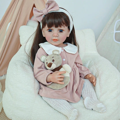 Reborn toddler doll in a pink dress, seated on a chair with a plush toy, featuring a large bow in her hair.