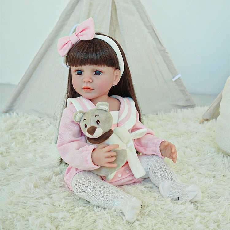 Reborn toddler doll with long brown hair, dressed in a pink and white outfit, holding a teddy bear.