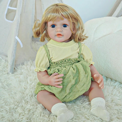 Reborn toddler doll sitting on a rug, wearing a green outfit with a light green top and white socks.