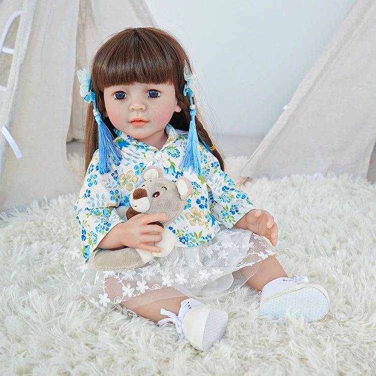 Reborn toddler doll sitting on a soft rug, wearing a floral blue and white top and holding a small teddy bear.