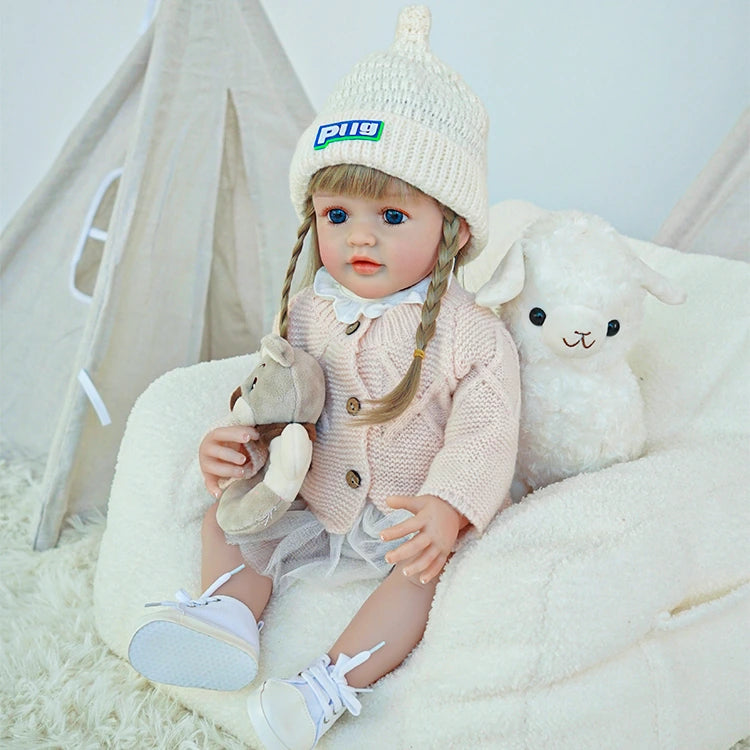 Reborn toddler doll sitting next to a plush toy, dressed in a pink outfit and white shoes.