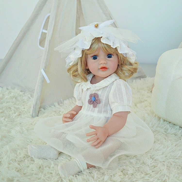 Reborn toddler doll sitting on a soft rug, dressed in a white outfit with a bonnet, holding her hands together.