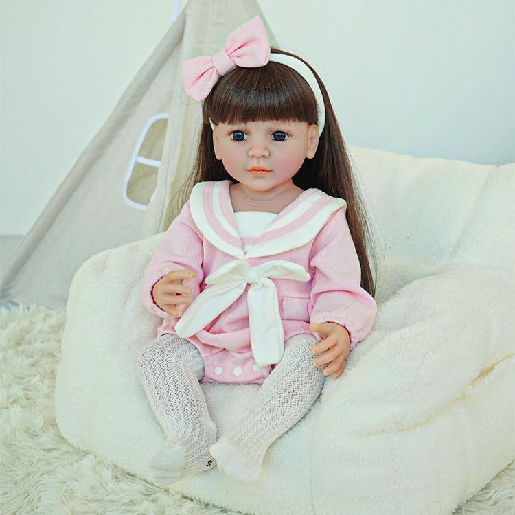 Reborn toddler doll sitting comfortably on a soft chair, dressed in a pink outfit with a large white bow and gray tights.