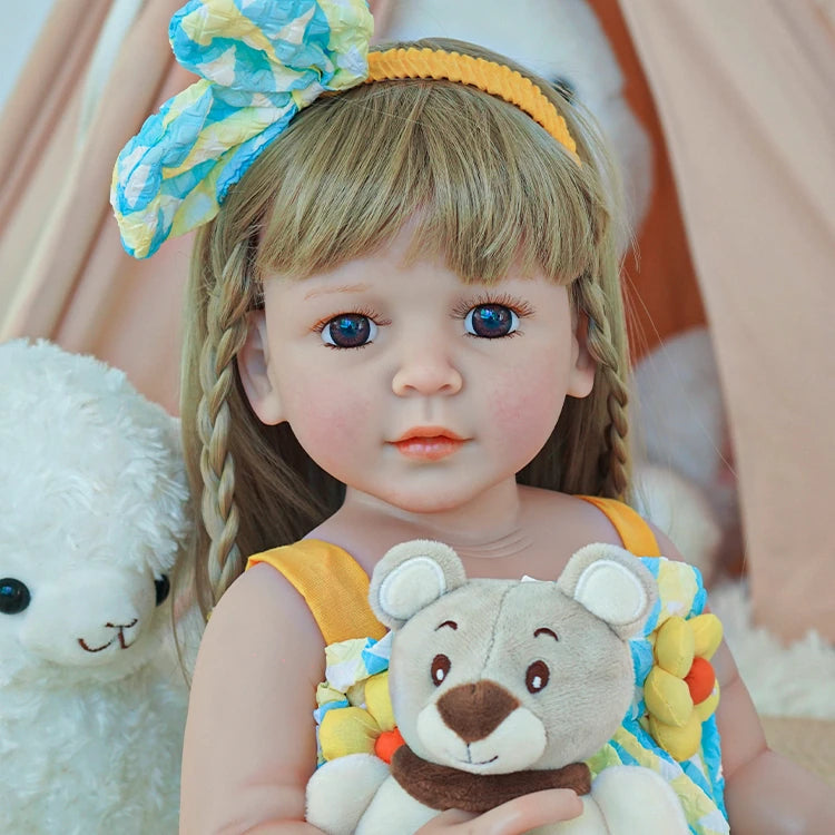 Close-up of reborn toddler doll with blonde hair and a blue bow, dressed in a yellow and blue outfit, holding a teddy bear.