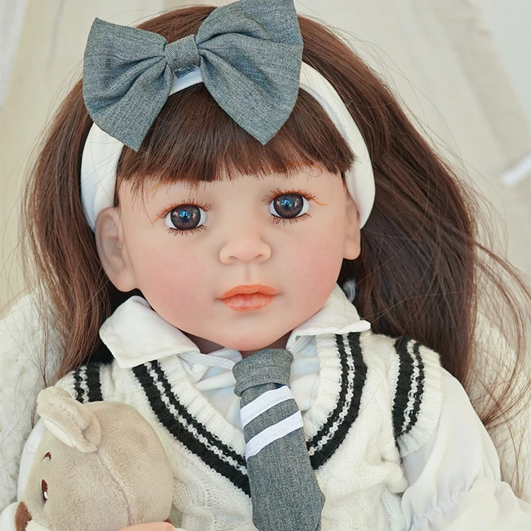 Close-up of reborn toddler doll with a large gray bow in her hair, wearing a white shirt and gray tie, holding a teddy bear.