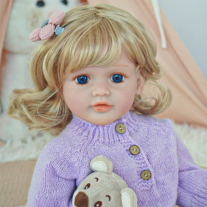 Close-up of reborn toddler doll with blonde hair and a pink bow, dressed in a purple sweater and holding a teddy bear.