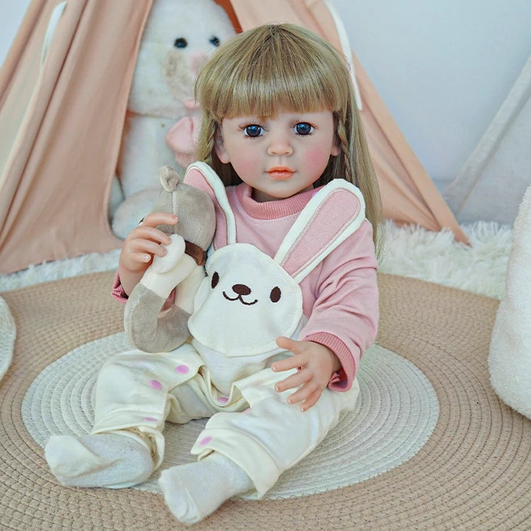 Reborn toddler doll sitting on a rug, dressed in white overalls with a bunny face and holding a small plush toy.