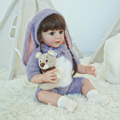 Reborn toddler doll sitting in a cute bunny-themed outfit with a hood, holding a plush bear on her lap.