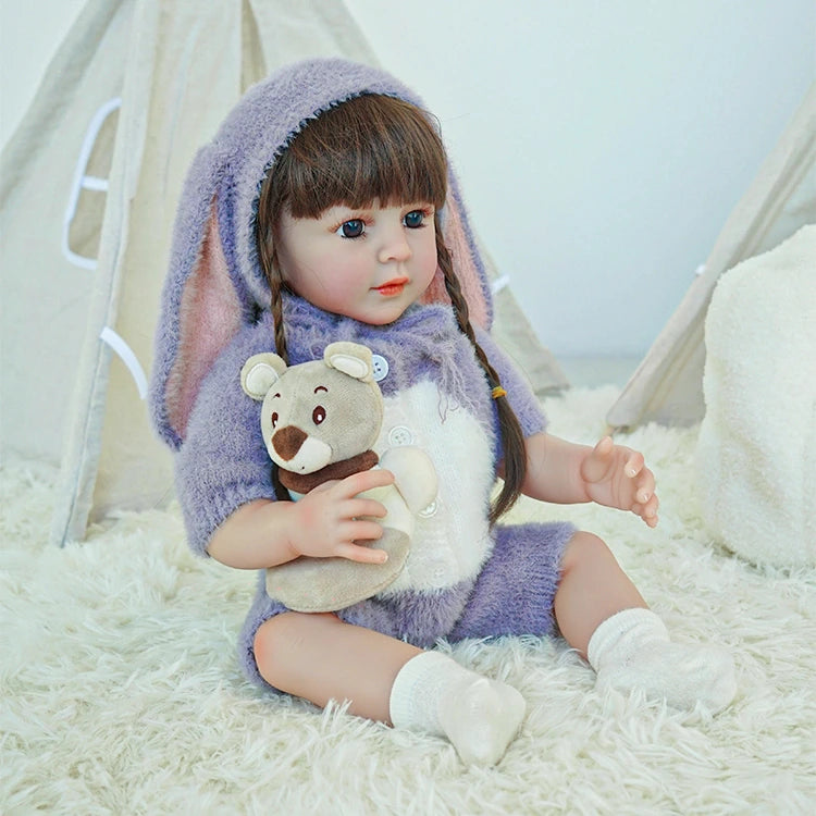 Reborn toddler doll sitting in a cute bunny-themed outfit with a hood, holding a plush bear on her lap.