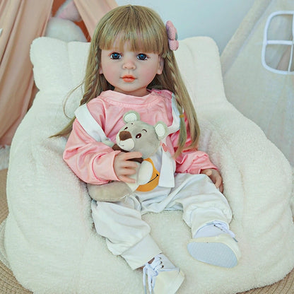 Reborn toddler doll sitting on a plush chair, wearing a pink outfit with white overalls, holding a teddy bear.