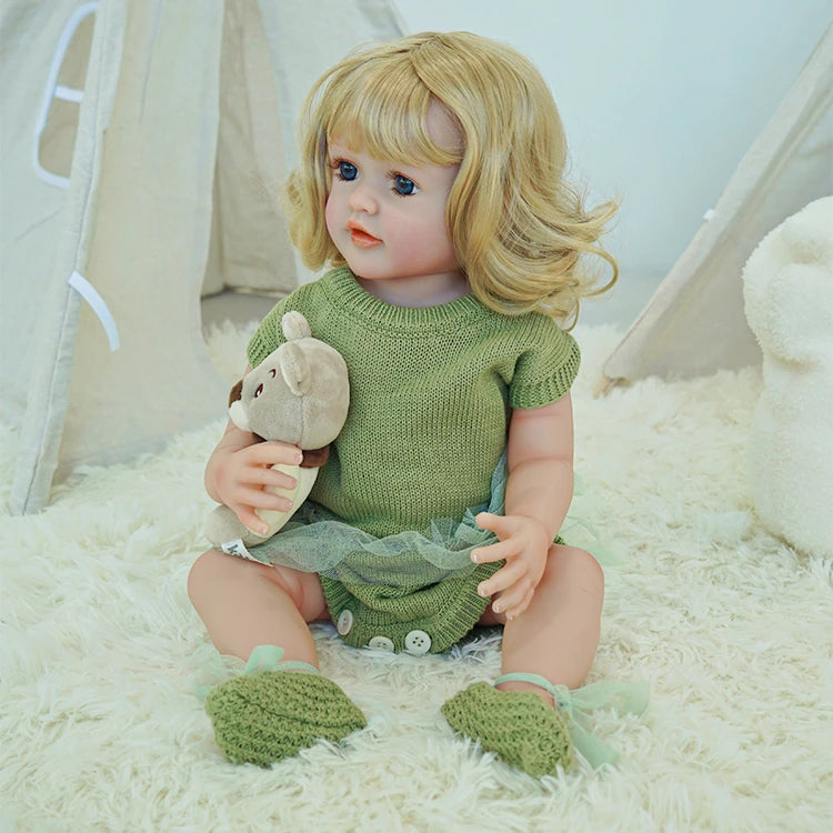 Reborn toddler doll in a green outfit, sitting beside a pair of knit shoes and holding a plush toy.