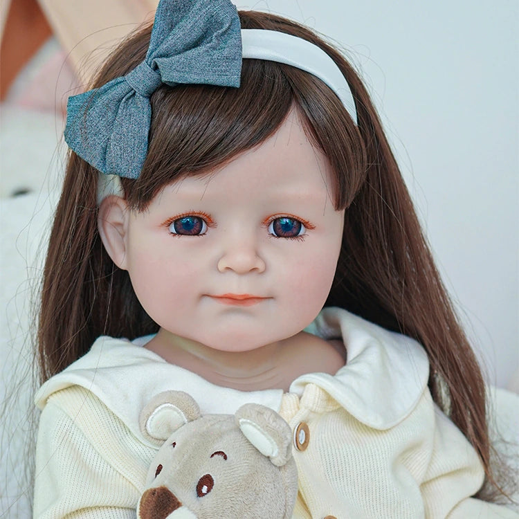 Close-up of reborn toddler doll with long brown hair and a large blue bow, wearing a cream sweater and holding a plush bear.