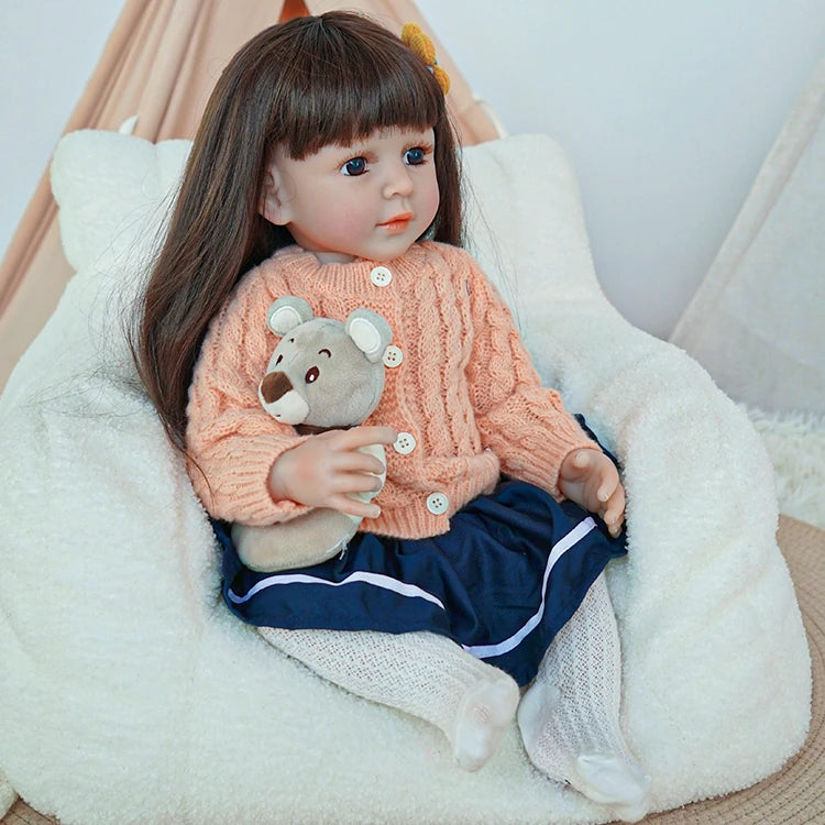 Reborn toddler doll sitting comfortably in a chair, dressed in a peach knit sweater and holding a teddy bear.