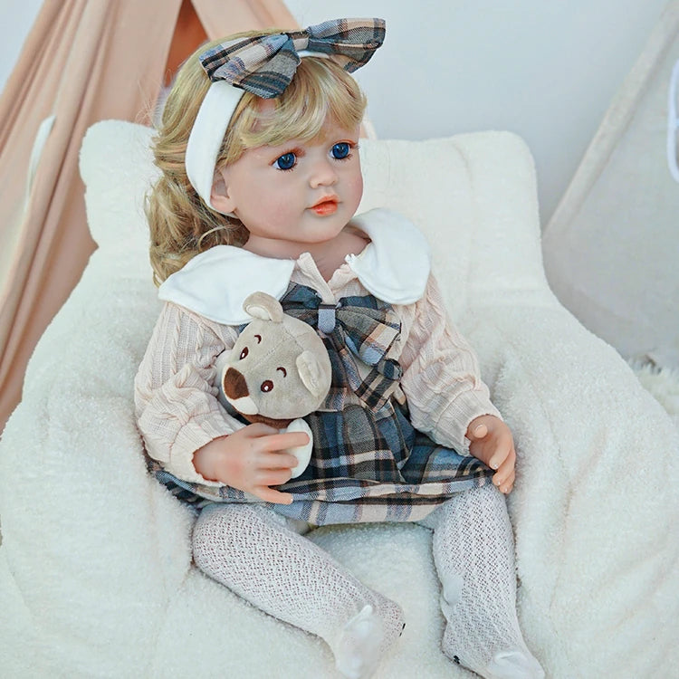 Reborn toddler doll sitting in a plush chair, dressed in a plaid dress with a large white collar and white tights, holding a plush teddy bear.