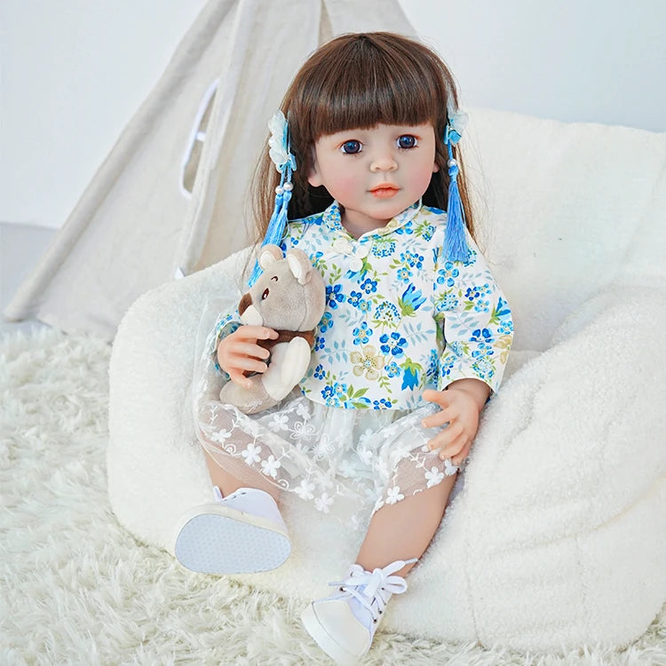 Reborn toddler doll sitting in a plush chair, dressed in a blue floral top and white skirt, holding a teddy bear.