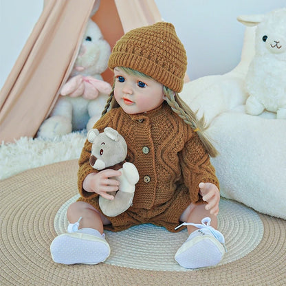 Reborn toddler doll wearing a cozy brown outfit, seated on a soft rug and holding a plush toy.