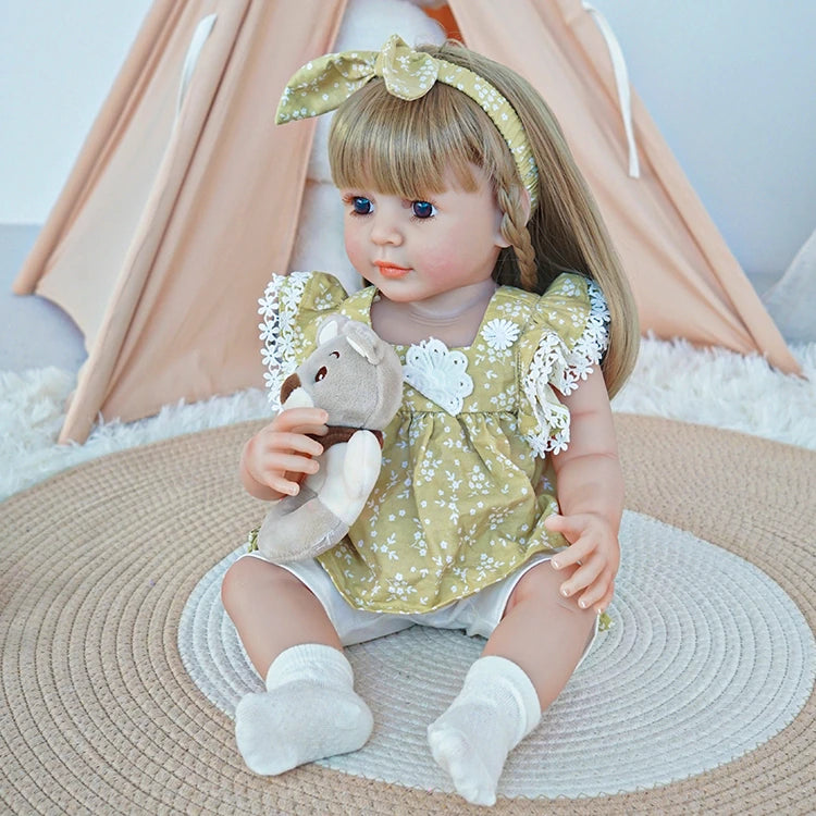 Reborn toddler doll sitting on a rug, dressed in a green floral dress with white socks, holding a plush toy, with her hair styled in bangs and a bow.