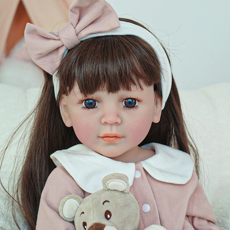 Close-up of a reborn toddler doll with straight brown hair, wearing a pink dress and holding a teddy bear.