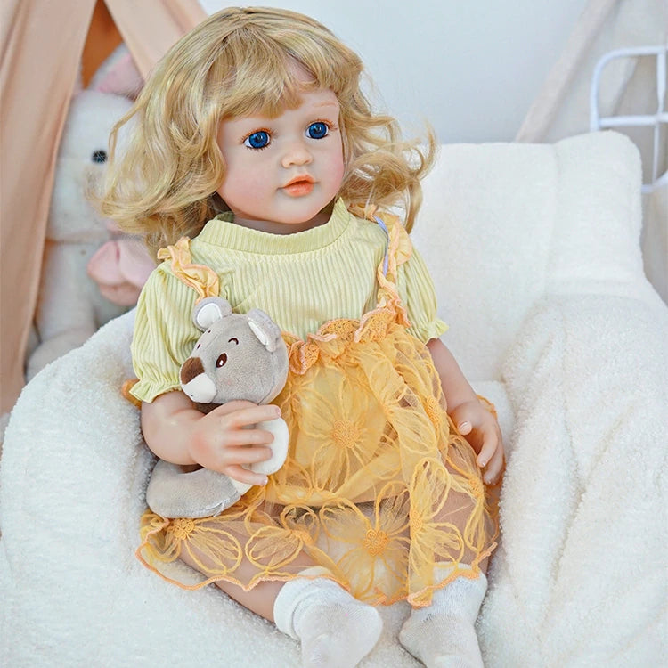 Reborn toddler doll sitting comfortably in a chair, dressed in a yellow dress with floral details and white socks, holding a teddy bear.
