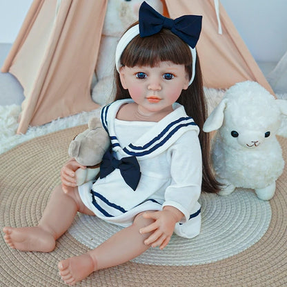 Reborn toddler doll sitting on a rug, dressed in a white and navy sailor outfit, holding a teddy bear and gazing to the side.