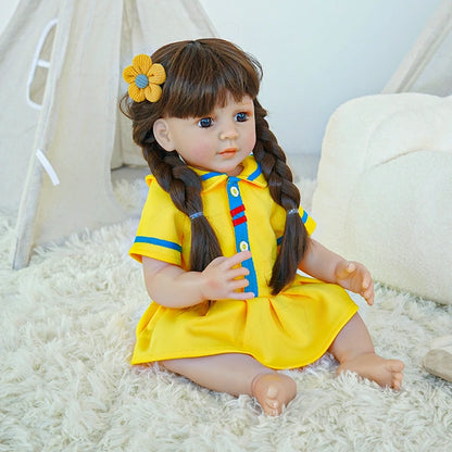 Reborn toddler doll sitting on a rug, dressed in a yellow dress with blue buttons, holding a plush toy, with her hair styled in two braids.