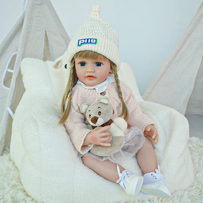 A reborn toddler doll holding a stuffed bear, wearing a cozy outfit with white shoes.