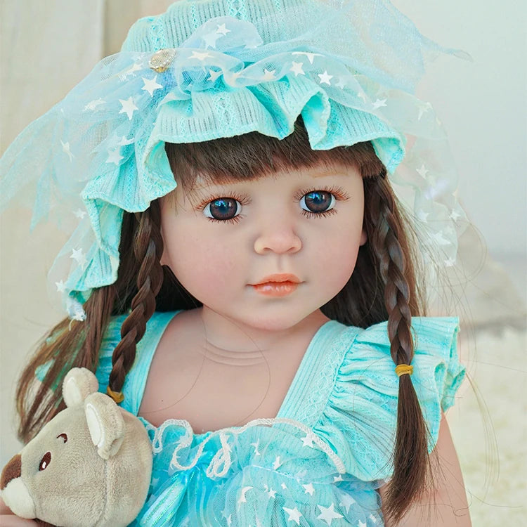 Close-up of reborn toddler doll wearing a light blue bonnet and dress, holding a plush teddy bear.