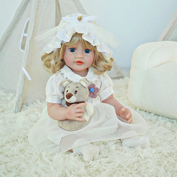 Reborn toddler doll sitting on a plush surface, wearing a white dress and bonnet, holding a small teddy bear.