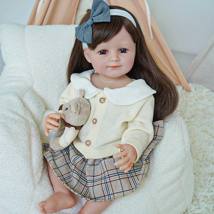  Reborn toddler doll sitting on a soft chair, dressed in a cream cardigan and plaid skirt, holding a small teddy bear.