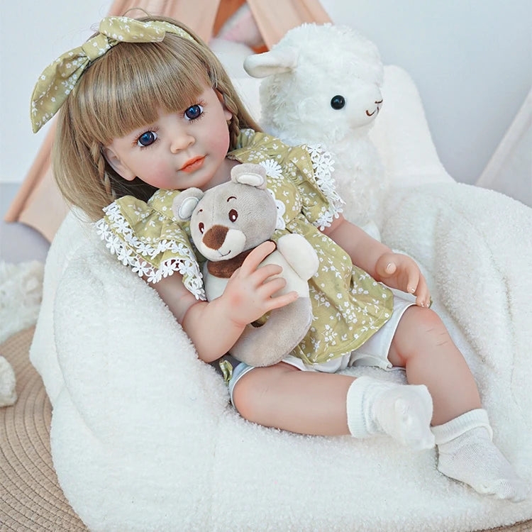 Reborn toddler doll sitting on a plush chair, dressed in a green floral dress, holding a teddy bear, looking forward.