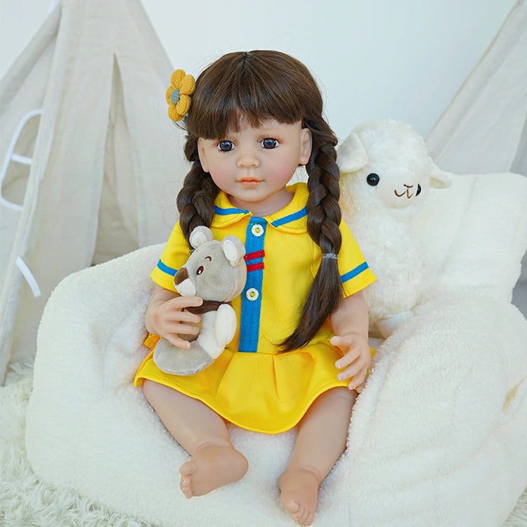 Reborn toddler doll sitting on a plush chair, dressed in a yellow and blue dress, holding a teddy bear, looking forward.