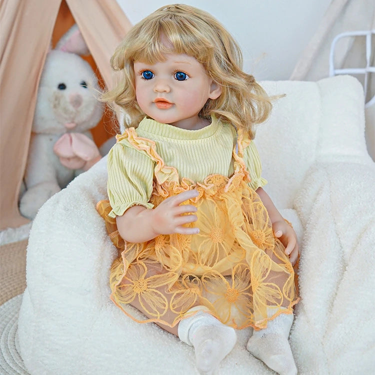 Reborn toddler doll sitting in a plush chair, dressed in a yellow floral dress, holding a teddy bear, looking forward.