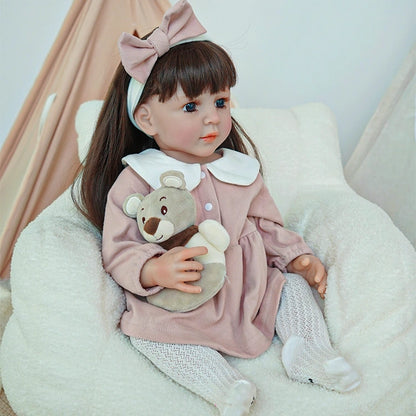 Reborn toddler doll sitting on a soft chair, dressed in pink with white socks, holding a small plush bear.