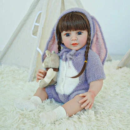 Reborn toddler doll in a soft purple bunny outfit with a hood and long bunny ears, sitting on a fluffy surface.