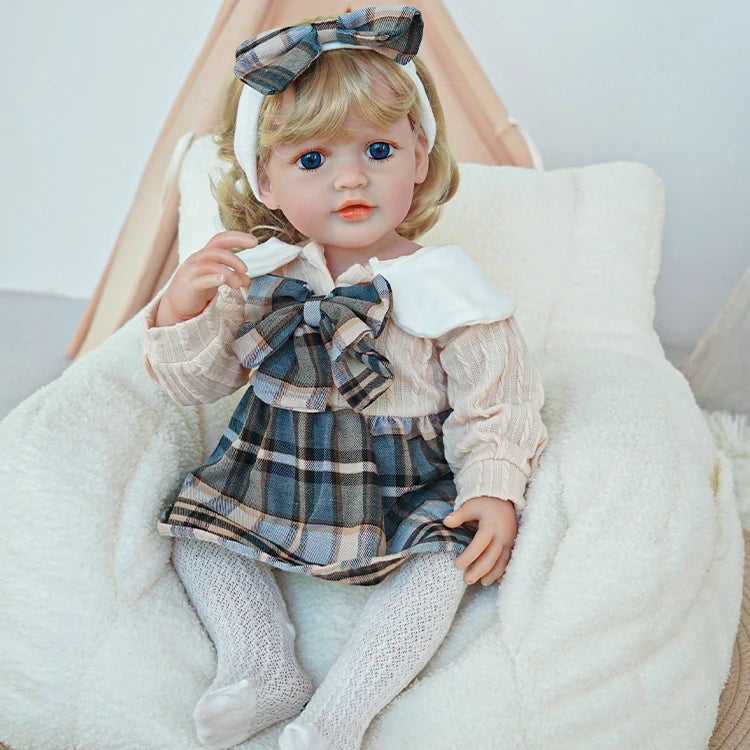 Reborn toddler doll sitting in a soft chair, dressed in a plaid dress and holding a teddy bear, looking gently to the side.